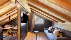 A cozy attic room with a wooden ceiling, a gray sofa, a small round table, and a TV mounted on the wall. - Chalet 'Rain Villa Genf' with Mountain View, Shared Garden and Wi-Fi (Livigno)