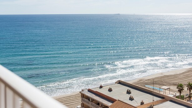 Un hotel frente al mar con terraza en la azotea y vistas a la playa de arena, bajo un cielo despejado.