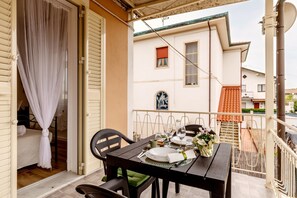 Een balkon met een eettafel gedekt voor twee personen, een stoel, een klein tafeltje met een bloevas en uitzicht op een gebouw met een rood dak.