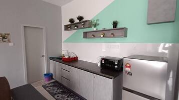 A compact kitchen with a refrigerator, microwave, and a small countertop. There are shelves above the counter holding decorative items and plants.