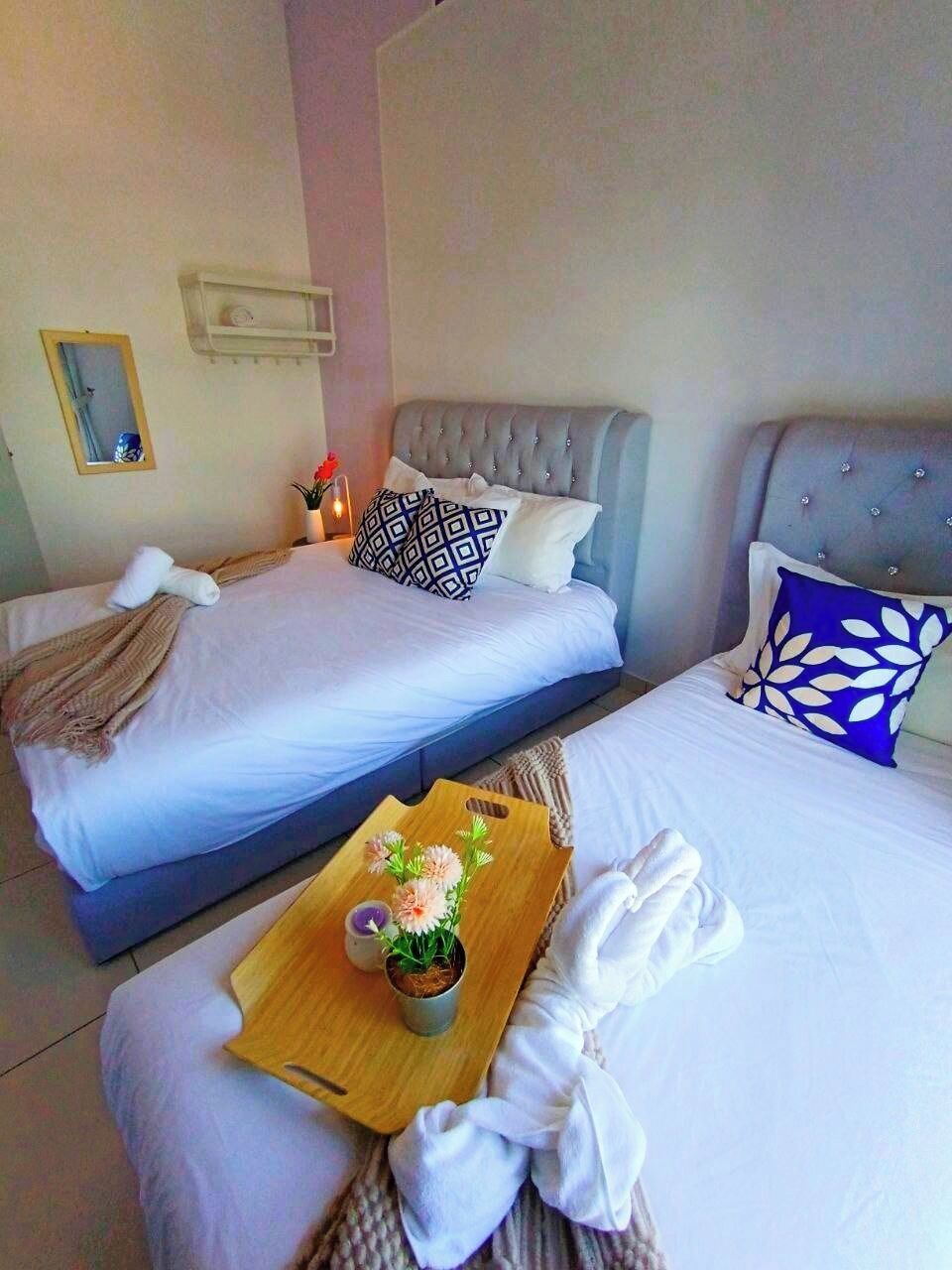 A hotel room with two beds, each with a tufted headboard, white linens, and decorative pillows. A wooden tray with flowers and a candle is placed on the bed.