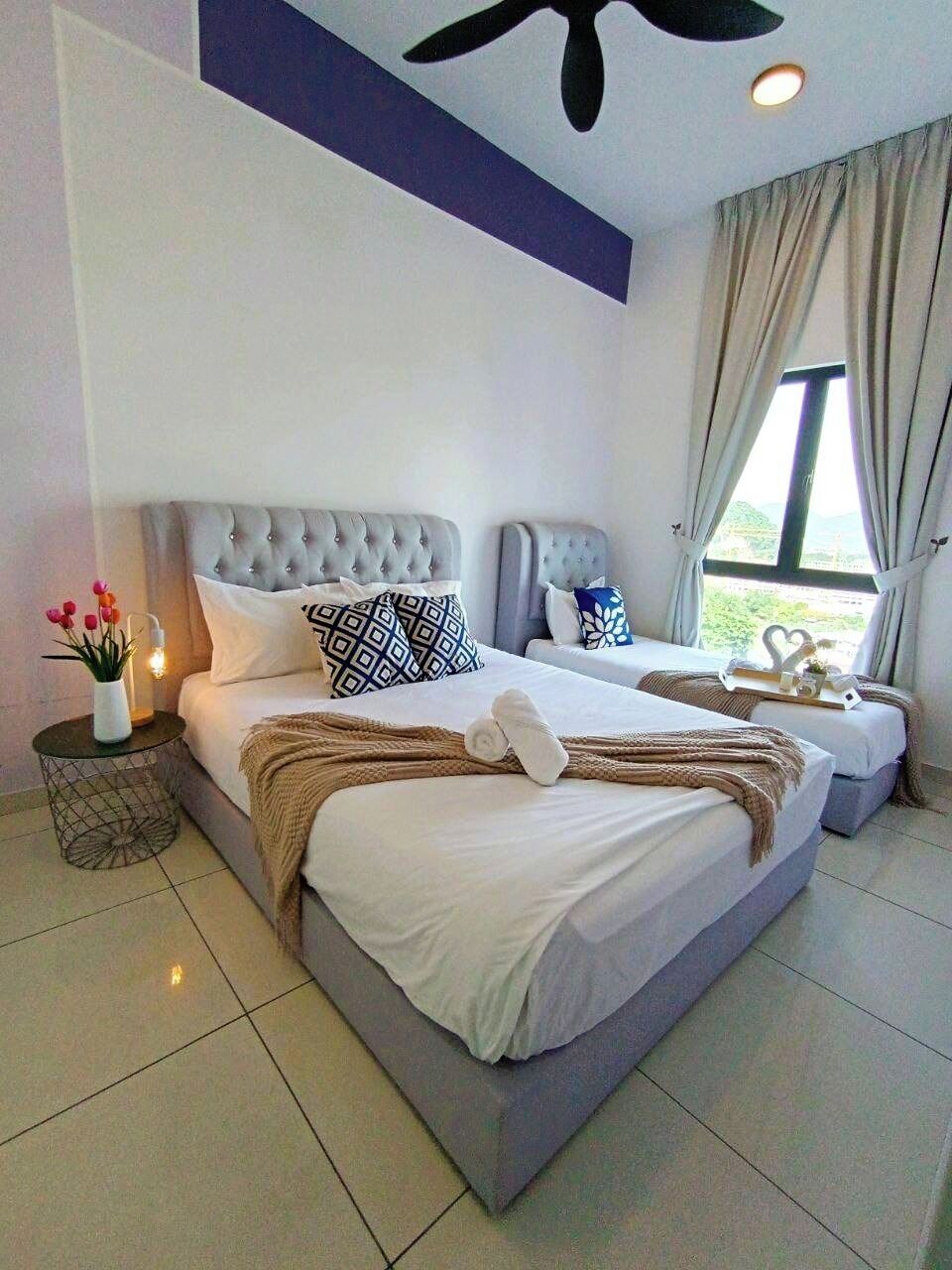 A double bed with a tufted headboard, a bedside table with a vase and a lamp, and a window with curtains.