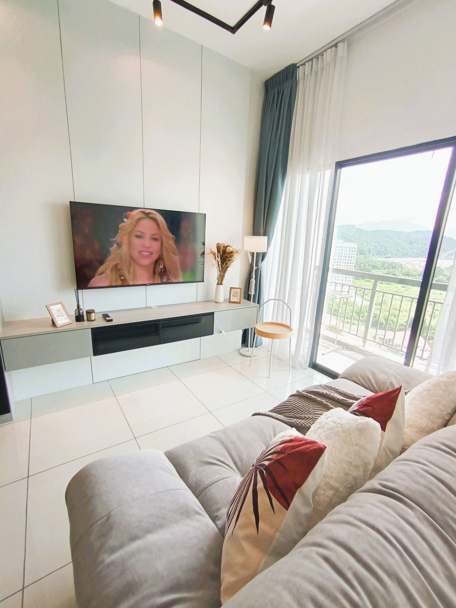 Un salon moderne avec un téléviseur à écran plat fixé au mur, un canapé agrémenté de coussins décoratifs, une table d’appoint avec une lampe, et une vue sur la verdure grâce à de grandes fenêtres.