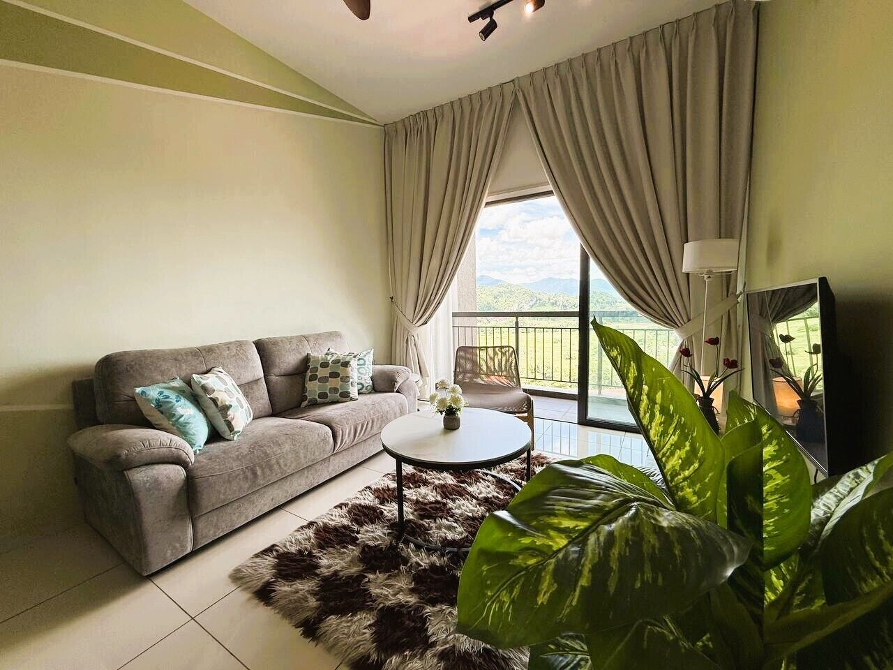 A living room with a sofa, a round coffee table, a large plant, and a balcony with curtains.