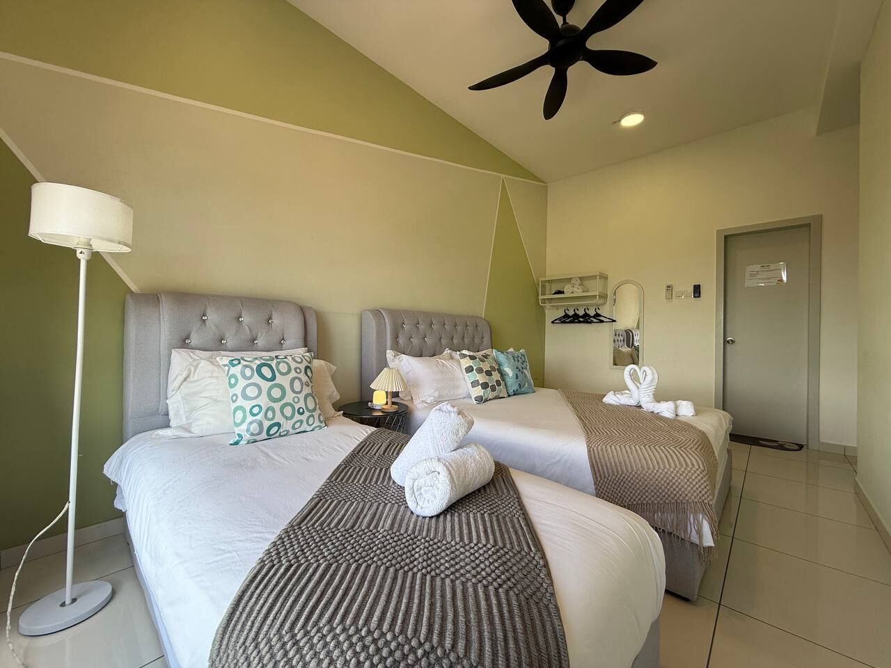 A hotel room with two beds, a ceiling fan, a bedside table with a lamp, and a door with a towel rack.