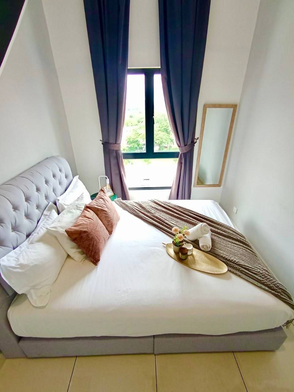 A neatly made bed with a tufted headboard, a bedside table with a bottle and a cup, and a view of greenery outside through a window with curtains.