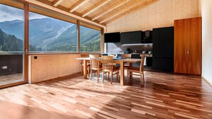 A spacious room with a large wooden dining table, chairs, a kitchen area, and a large window offering a mountain view. - Apartment 'Rumwaldhof Enzian' with Mountain View, Shared Terrace and Wi-Fi (Sulden)
