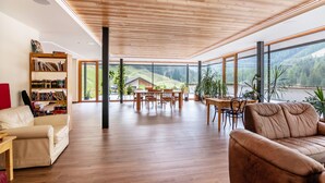 A spacious living area with a wooden ceiling, large windows, a dining table, and a bookshelf. - Apartment 'Rumwaldhof Enzian' with Mountain View, Shared Terrace and Wi-Fi (Sulden)