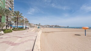 A beachside promenade with a sandy beach, palm trees, and a hotel building with balconies.