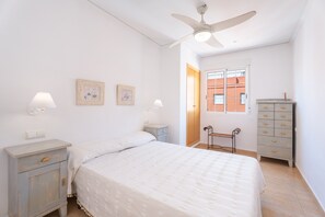 Une chambre à coucher comprenant un lit, deux tables de chevet, une commode, un ventilateur de plafond et une fenêtre donnant sur des immeubles.