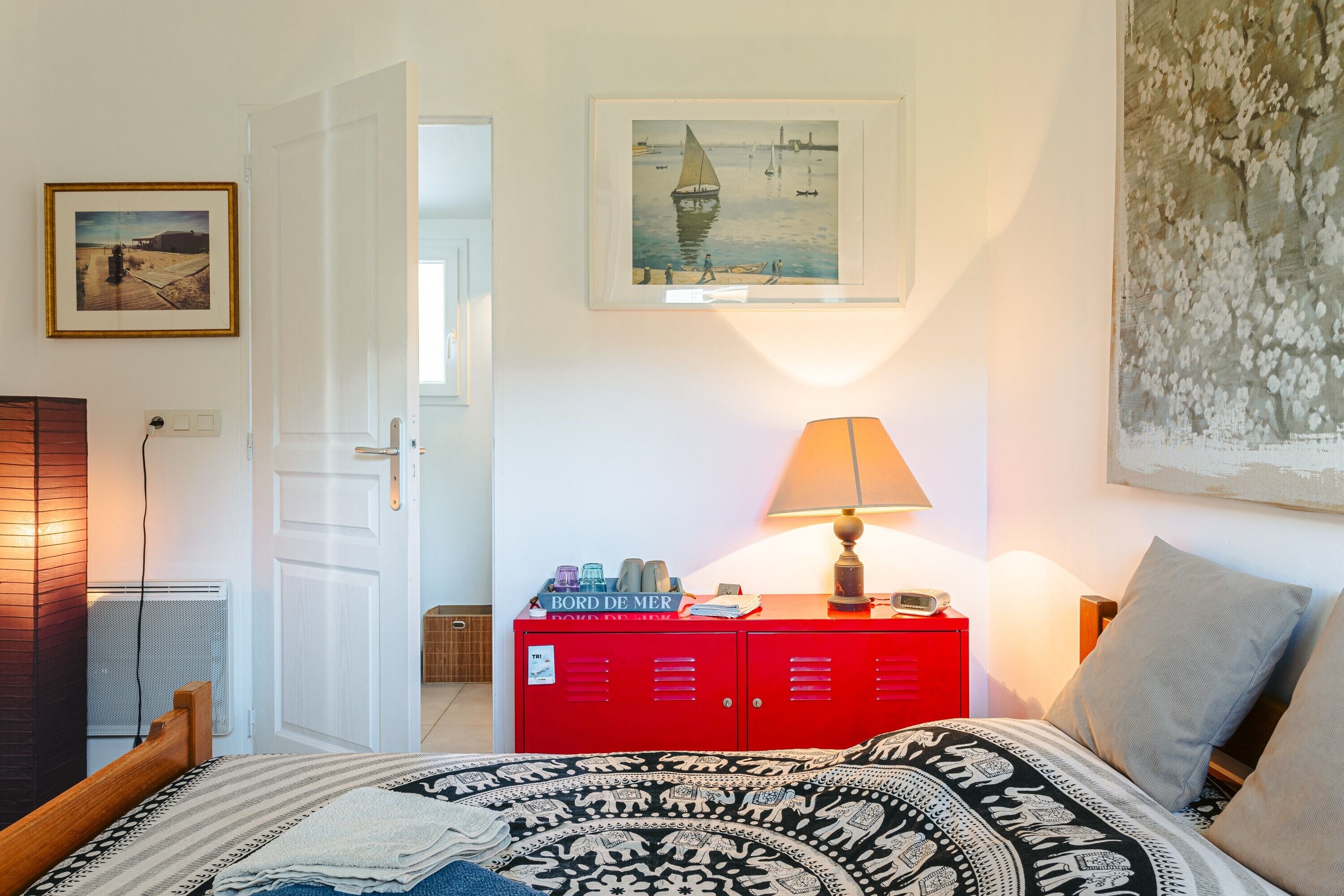 A bedroom with a bed, a red dresser, a lamp, and a painting on the wall.