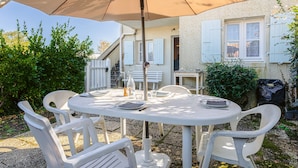 Een ronde buitentafel met witte stoelen, een beige parasol en een wit gebouw met luiken op de achtergrond.