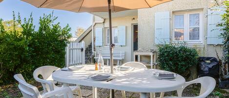 A round outdoor table with white chairs, a beige umbrella, and a white building with shutters in the background.