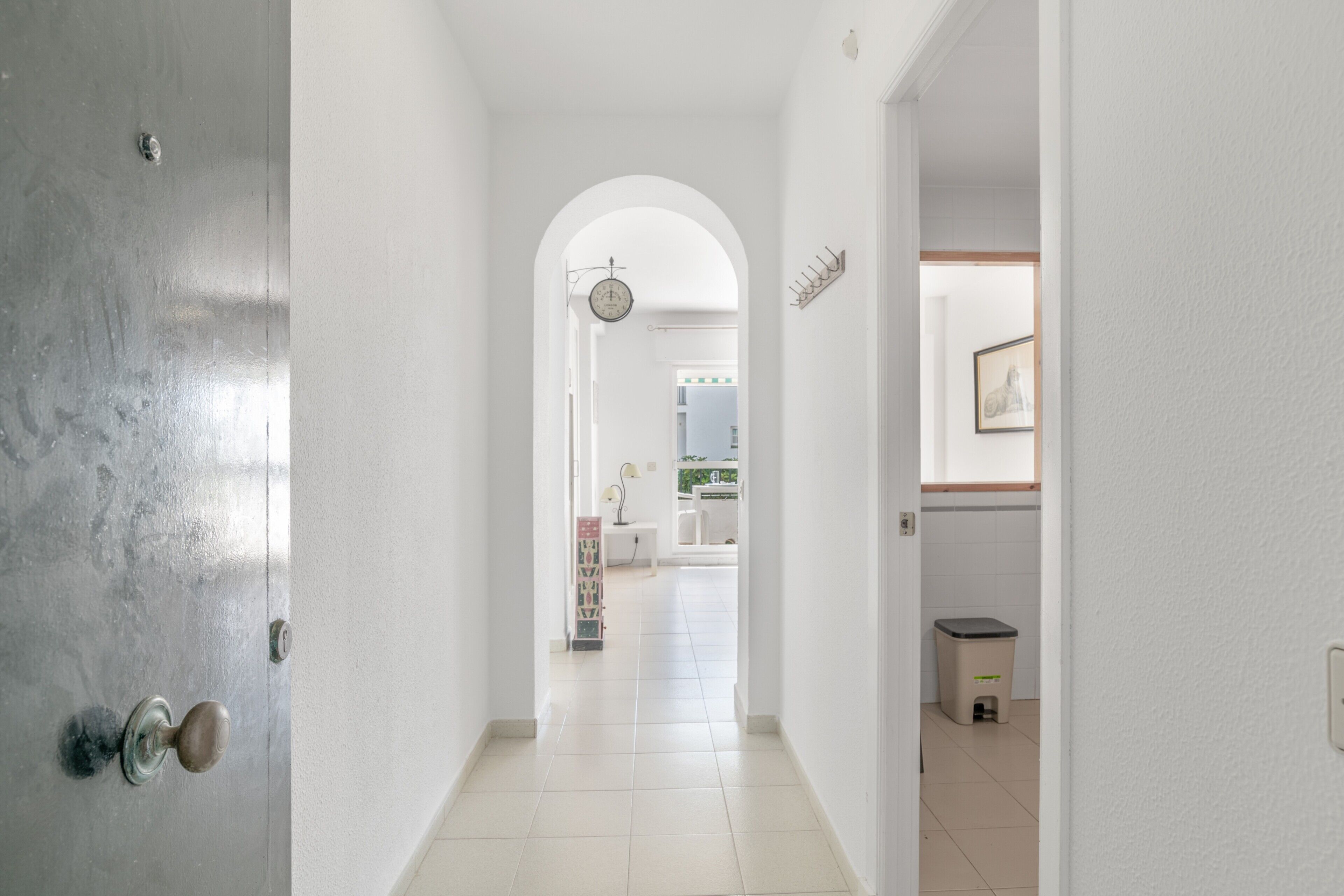 A hallway with a door, a clock, a trash bin, and a framed picture on the wall.
