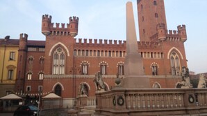 Un edificio histórico con una torre alta, un monumento central y estatuas.