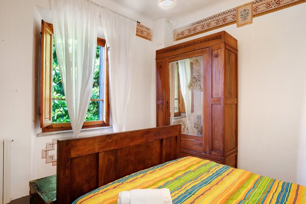 Un dormitorio con cama de madera, ventana con cortinas translúcidas, armario de madera y colcha estampada.