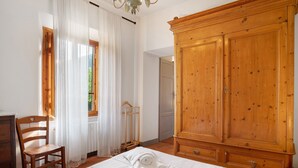 Une chambre avec une armoire en bois, un lit recouvert d’une couette blanche, une chaise en bois et une fenêtre ornée de rideaux légers.