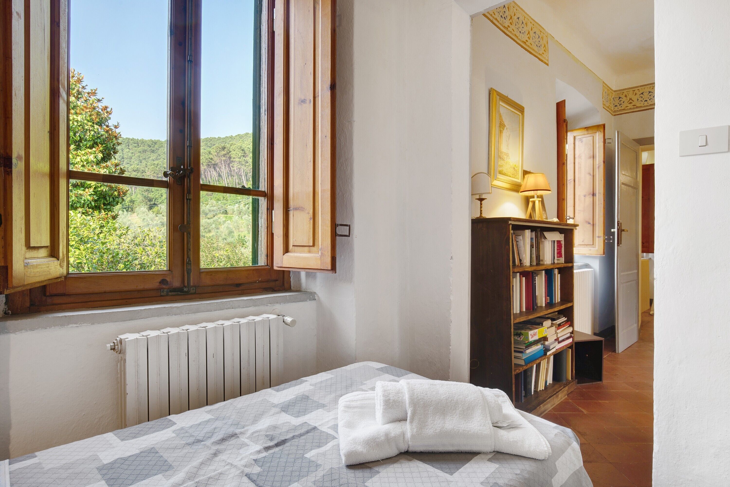 A bedroom with a bed, a radiator, a window with a view of greenery, a bookshelf, and a door leading to another room.