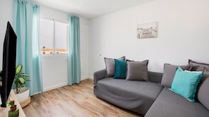 A living room with a grey sofa, teal cushions, a potted plant, and a painting on the wall. - Vacation Home 'Casa Alisios' with Mountain View, Private Terrace and Wi-Fi (Costa Calma)