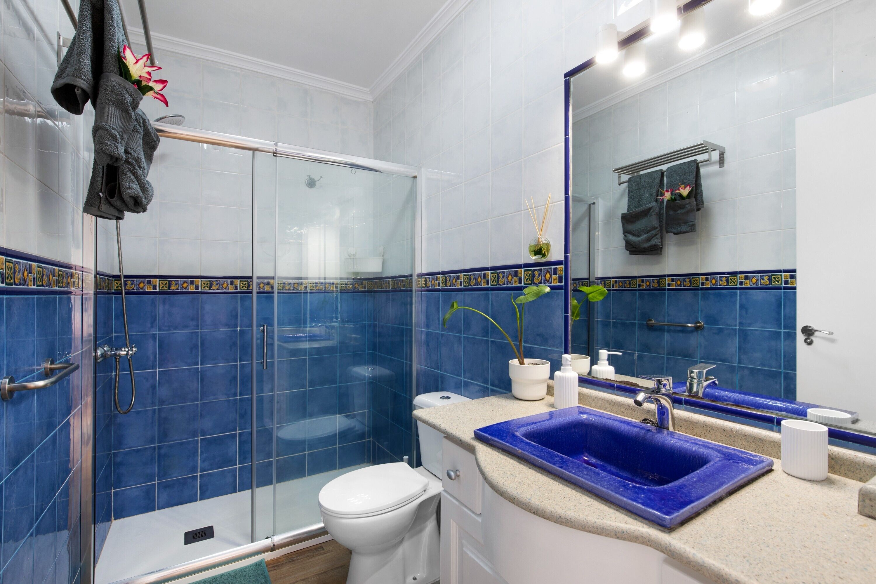 Ein Badezimmer mit Duschkabine aus Glas, Toilette in Weiß, Waschbecken in Blau und Spiegel mit Handtuchhalter.