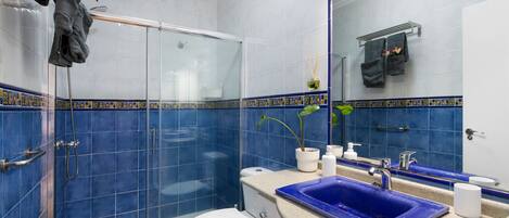 Ein Badezimmer mit Duschkabine aus Glas, Toilette in Weiß, Waschbecken in Blau und Spiegel mit Handtuchhalter.