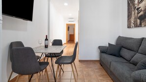 Een moderne woonkamer met een grijze bank, een eettafel met stoelen, een flatscreen-televisie aan de muur en een schilderij boven de bank.