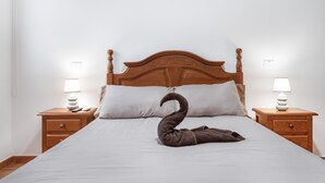 Una cama bien hecha con una toalla con forma de cisne, cabecero de madera, dos mesitas de noche con lámparas y una pared blanca lisa.