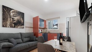 Una sala de estar moderna con un sofá gris, una mesa de comedor con dos sillas, un televisor montado en la pared y una cocina con armarios rojos y una ventana.