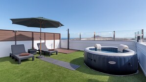Terraza en la azotea con césped artificial, sillas de descanso, una bañera de hidromasaje inflable y un paraguas grande.