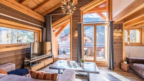 Interior de una cabaña de troncos moderna con televisor grande, asientos cómodos y ventanas amplias que ofrecen vistas a la montaña.