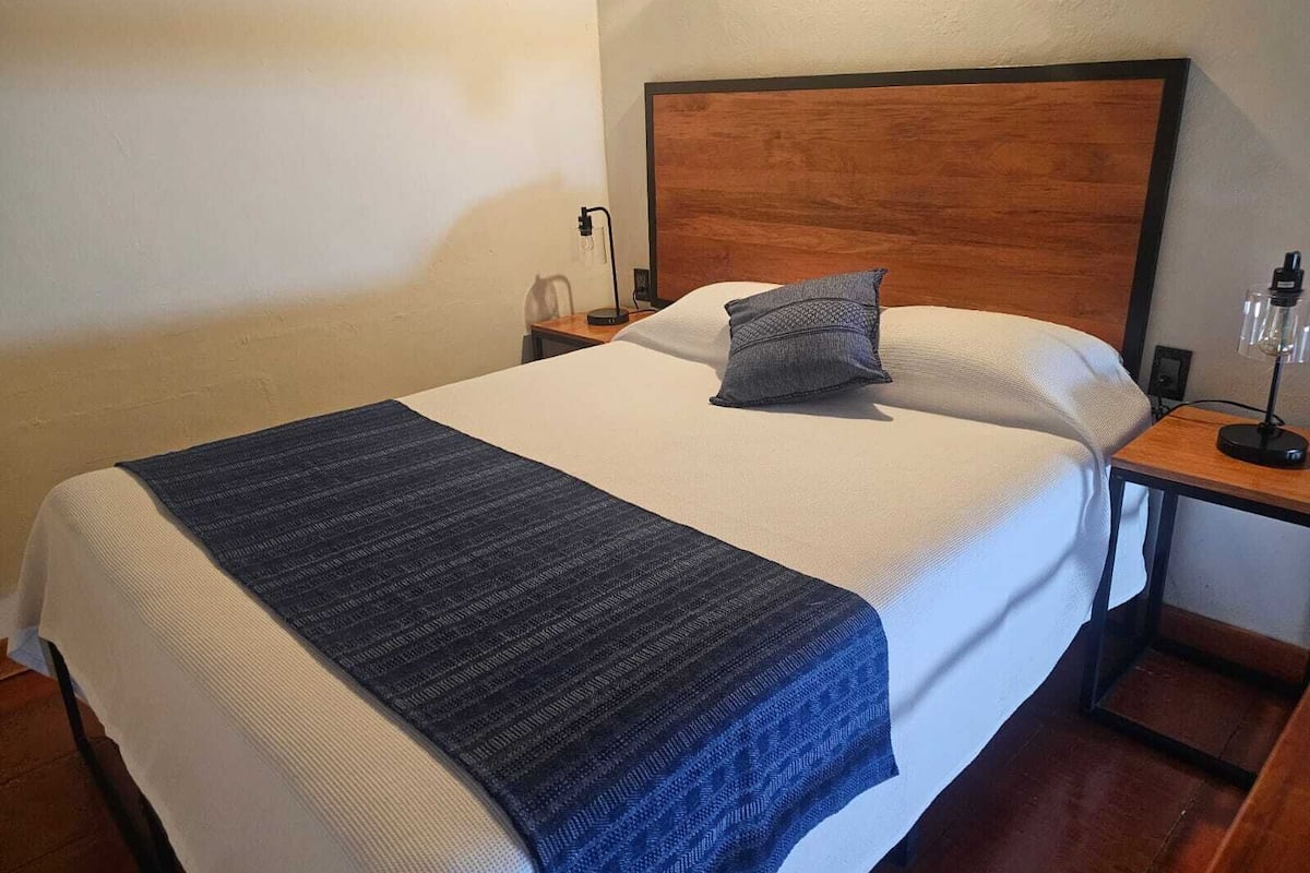 A neatly made bed with a blue blanket, a wooden headboard, a bedside lamp, and a nightstand with a phone and a lamp.