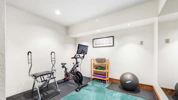 A small, well-lit room with a stationary bike, a stepper, a shelf with exercise balls, and a large exercise ball.