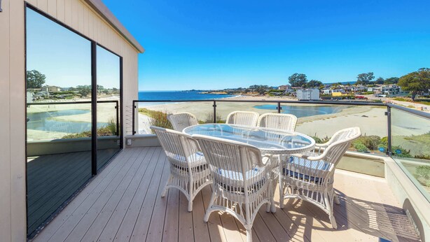 Un balcon avec une table en verre et des chaises en osier, donnant sur une plage et l’océan.