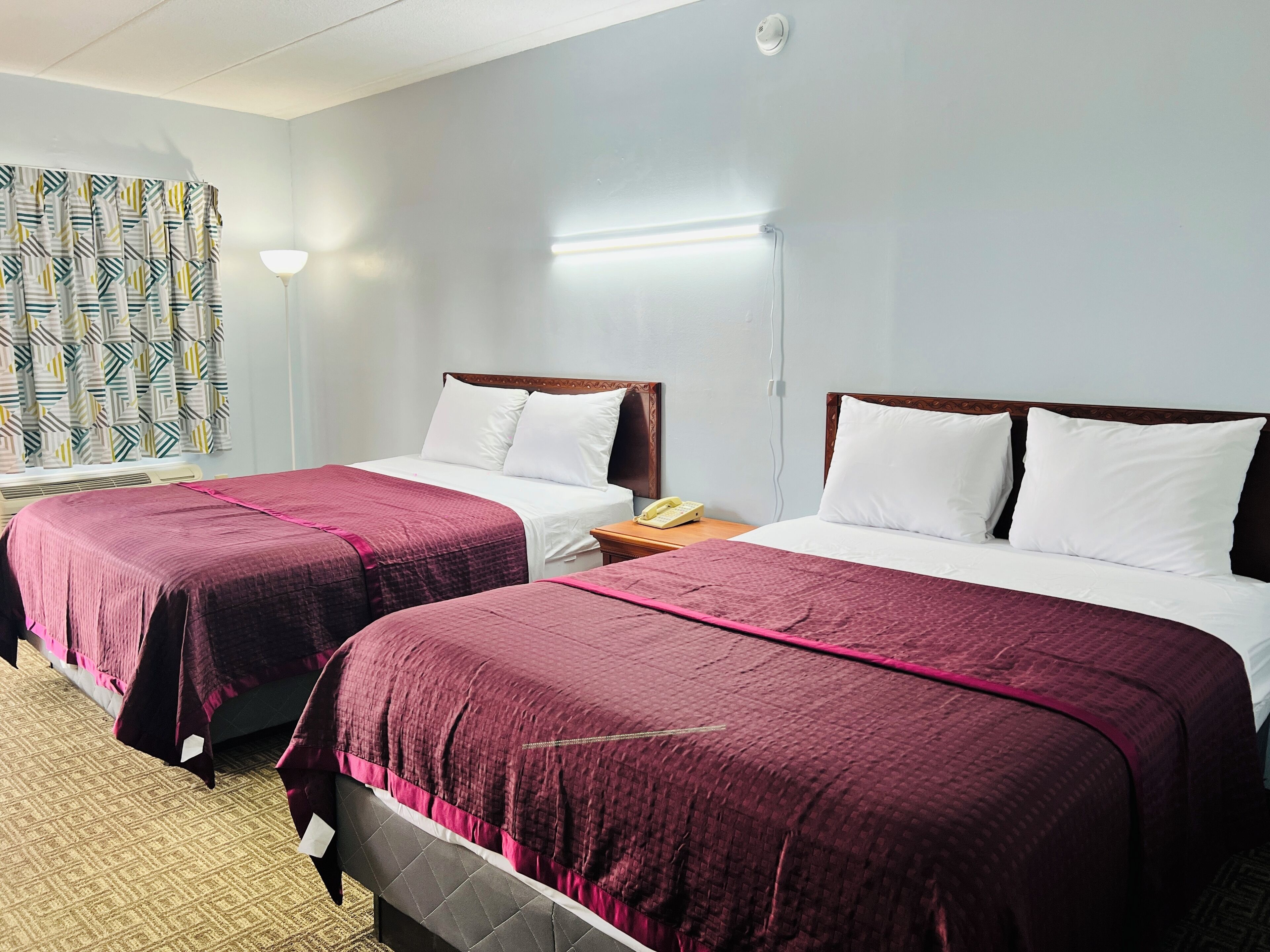 A hotel room with two beds, each with a wooden headboard, white bedding, and a pink accent. There is a bedside table with a lamp and a telephone on one side.