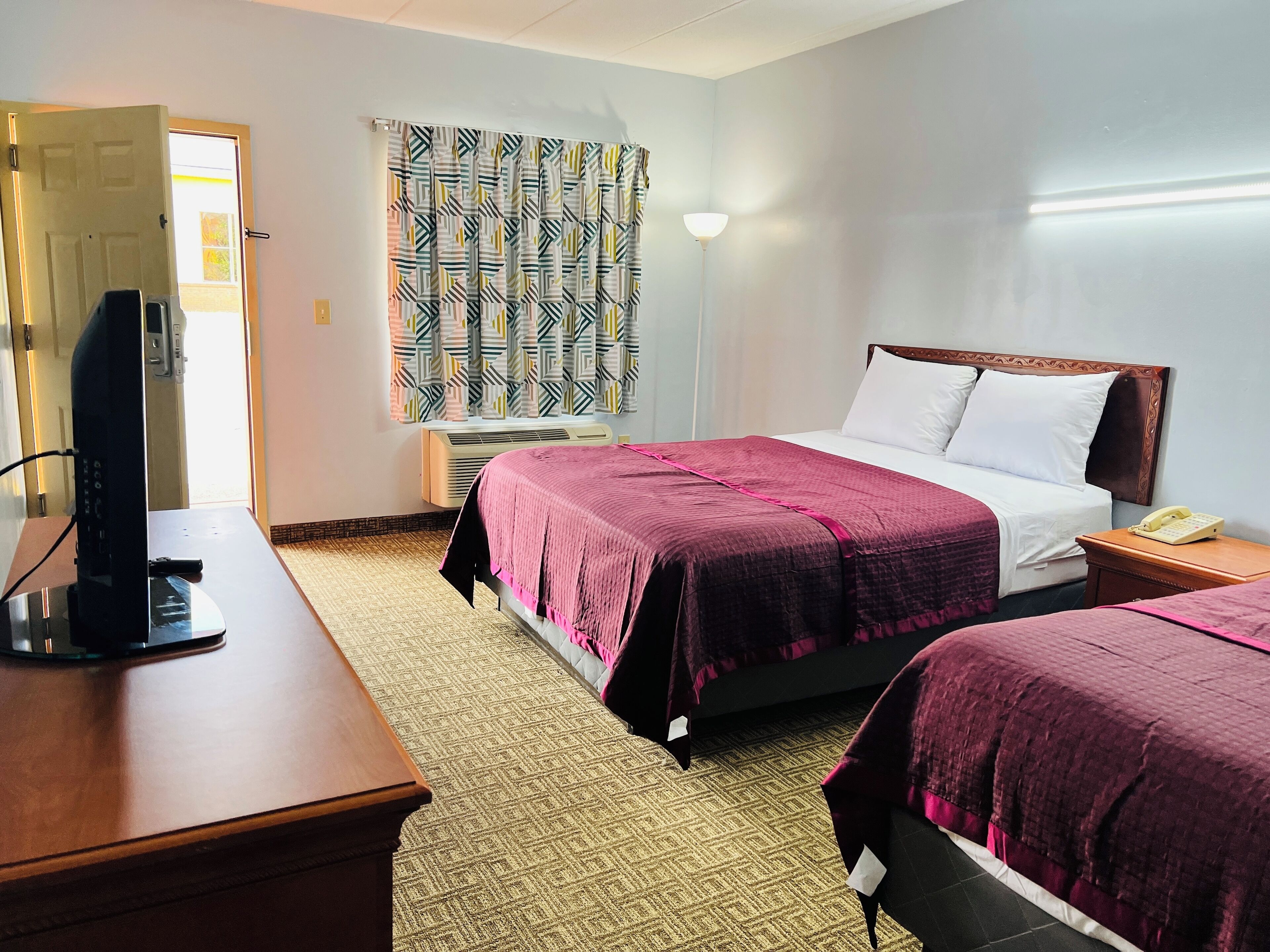 A hotel room with a bed, bedside table, TV, and a window with patterned curtains.