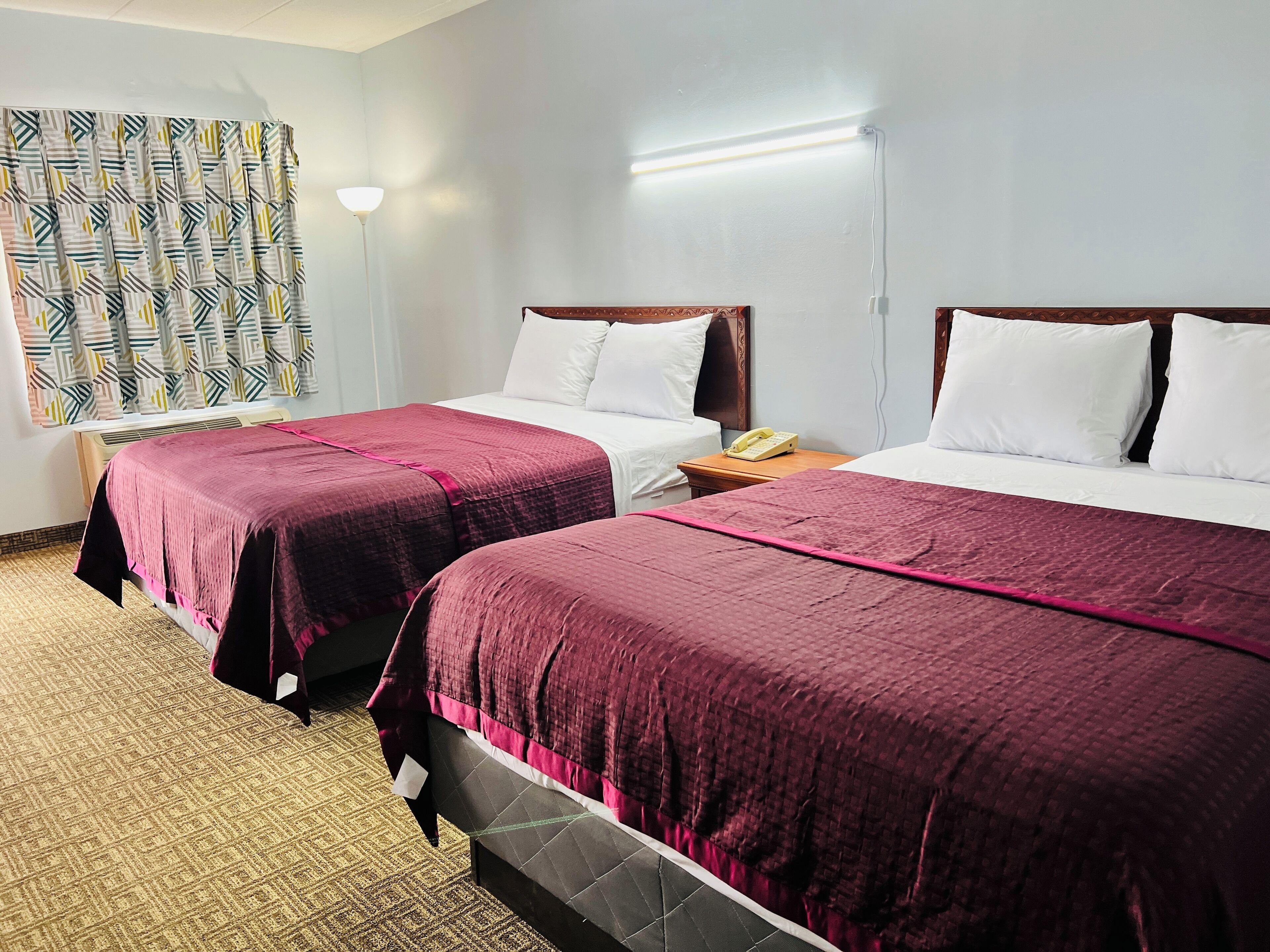 Ein Hotelzimmer mit zwei Betten, jeweils mit Kopfstütze, weißer Bettwäsche und einer roten Tagesdecke. Auf dem Nachttisch stehen eine Lampe und ein Telefon.