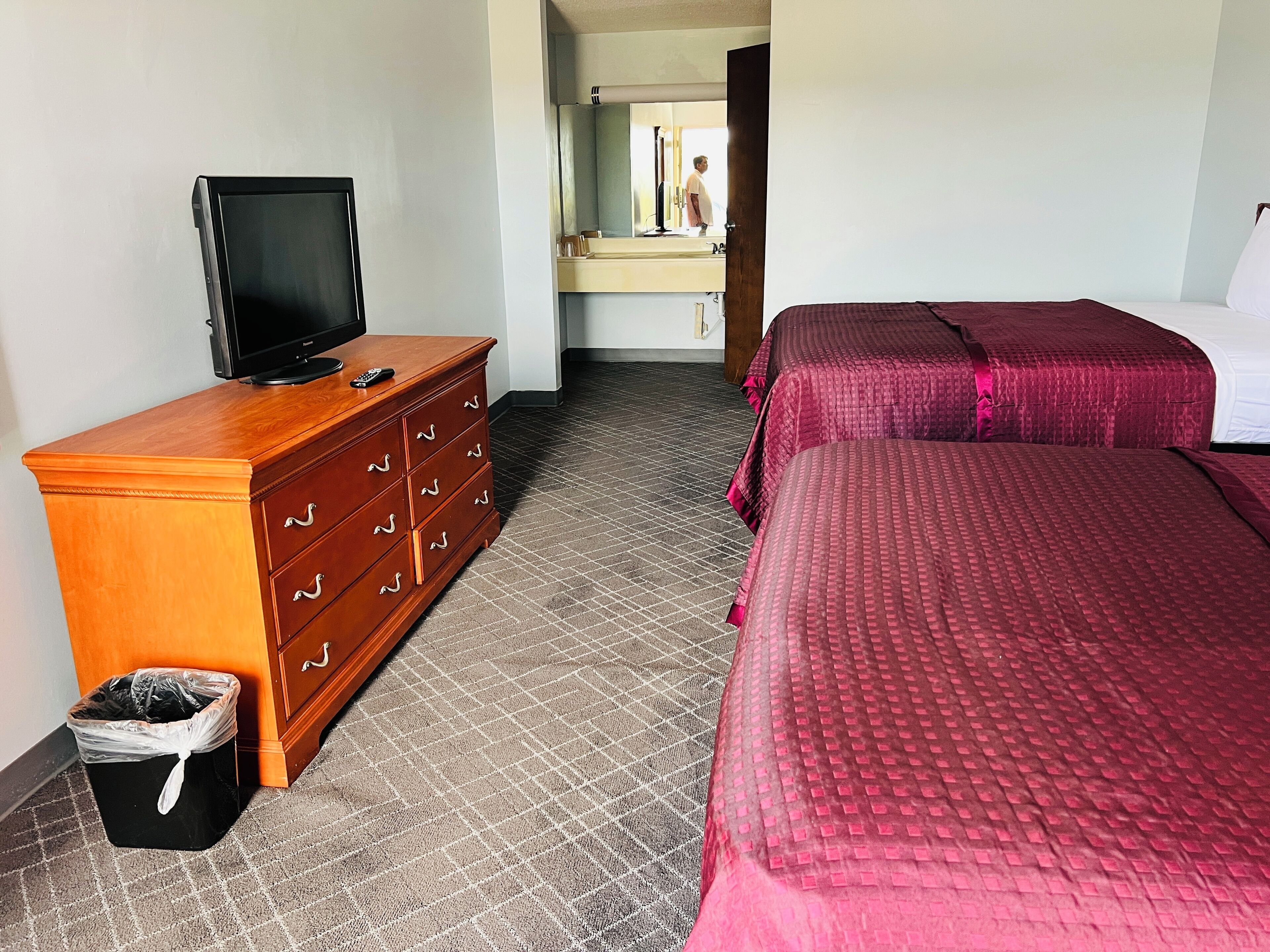 A hotel room with a wooden dresser, a television, a trash bin, and two beds with red bedding.