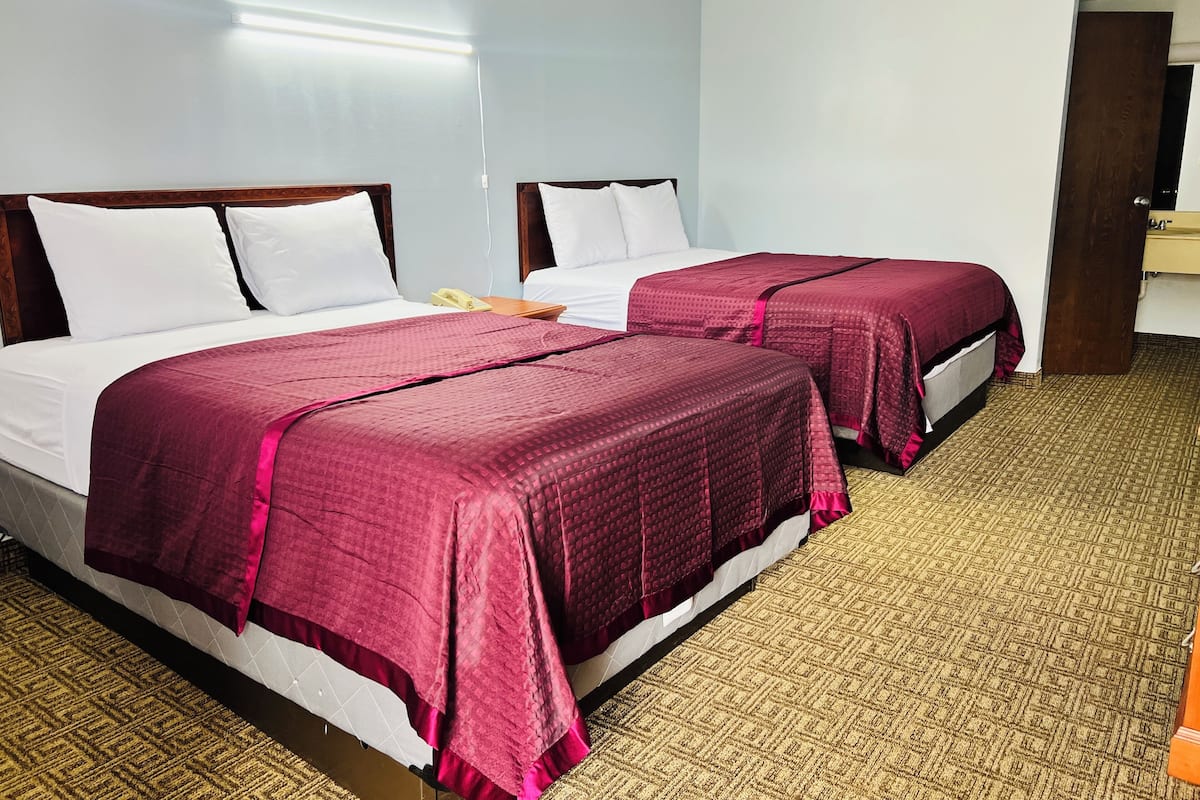 A hotel room with two beds, each with white bedding and maroon covers. There is a telephone on the nightstand, a wooden door, and a bathroom with a sink and mirror.