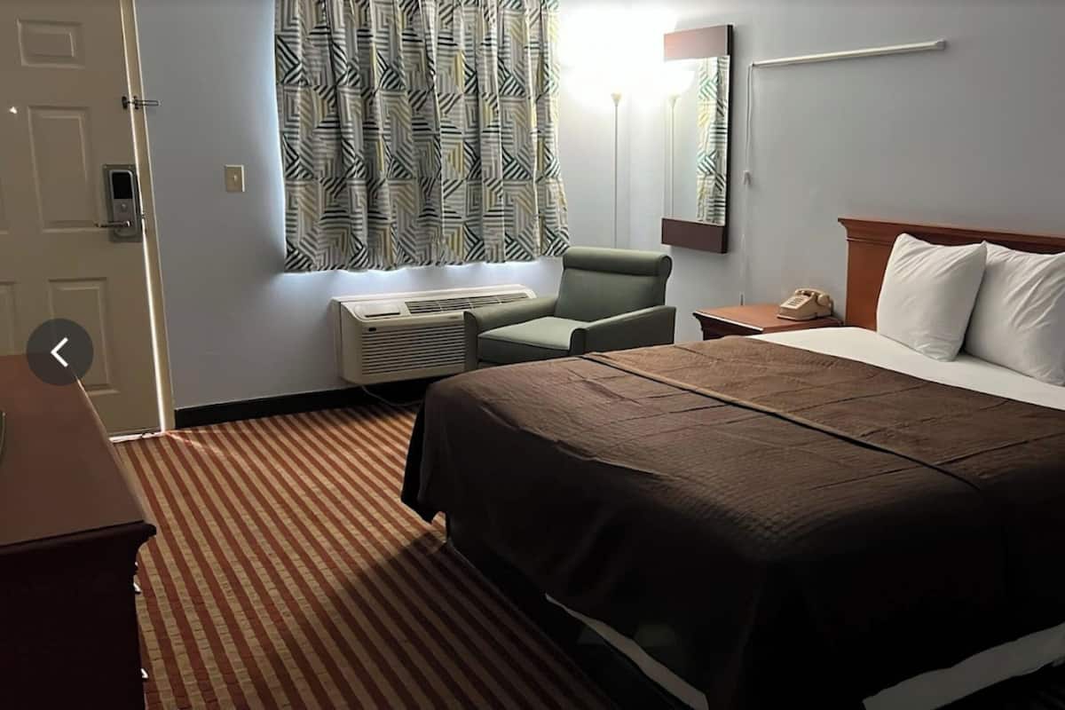 A hotel room with a large bed, a chair, an air conditioning unit, a bedside table with a phone, and a window with patterned curtains.
