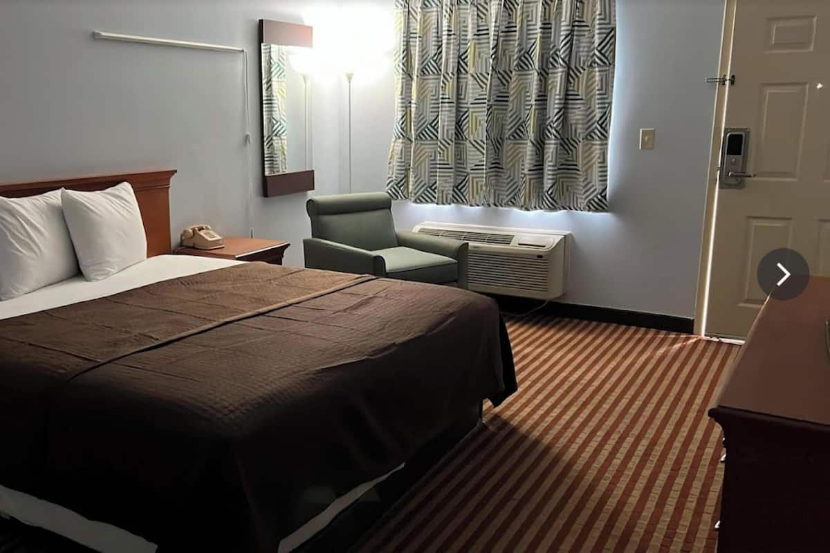 A hotel room with a large bed, a desk with a telephone, a chair, an air conditioning unit, and a patterned curtain.
