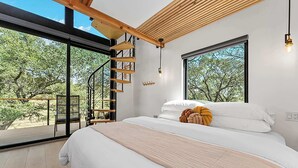 Un dormitorio con una cama grande, una escalera de madera y vistas a árboles a través de amplios ventanales.
