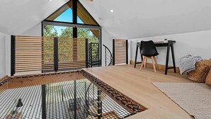 Un dormitorio tipo loft moderno con techo inclinado, ventanas amplias, piso de madera, una silla negra y una mesita.