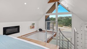 A bedroom with a sloped ceiling, a bed, a TV, a small shelf with a plant, and a view of the outdoors through a large window. - The Live Oak at Meridian, Hot tub & Firepit! (Fredericksburg)