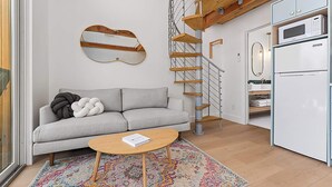 A modern living room with a grey sofa, a wooden coffee table, a colorful rug, a spiral staircase, and white cabinetry with a microwave and refrigerator. - The Live Oak at Meridian, Hot tub & Firepit! (Fredericksburg)