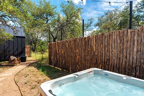 Un jardin avec un jacuzzi, une clôture en bois et une zone bordée d’arbres.