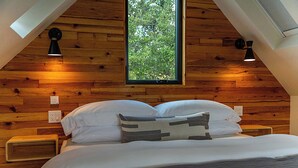 Ein Schlafzimmer mit schrägverdeckter Decke, Holzwänden, einem großen Fenster und zwei Nachttischlampen.