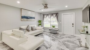 Una sala de estar moderna con un sofá blanco, un ventilador de techo, un ventanal con cortinas y un televisor de pantalla plana montado en la pared.