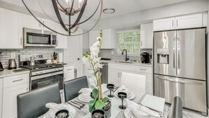 Una cocina moderna con armarios blancos, electrodomésticos de acero inoxidable, una mesa de comedor preparada para cuatro personas y una araña.