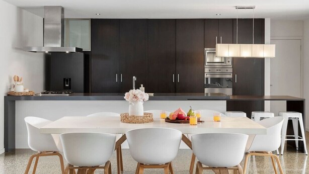 A modern kitchen with dark cabinetry, a central island, and a dining area with white chairs. - Luxury Beachside Entertainer With Heated Pool (St Leonards)