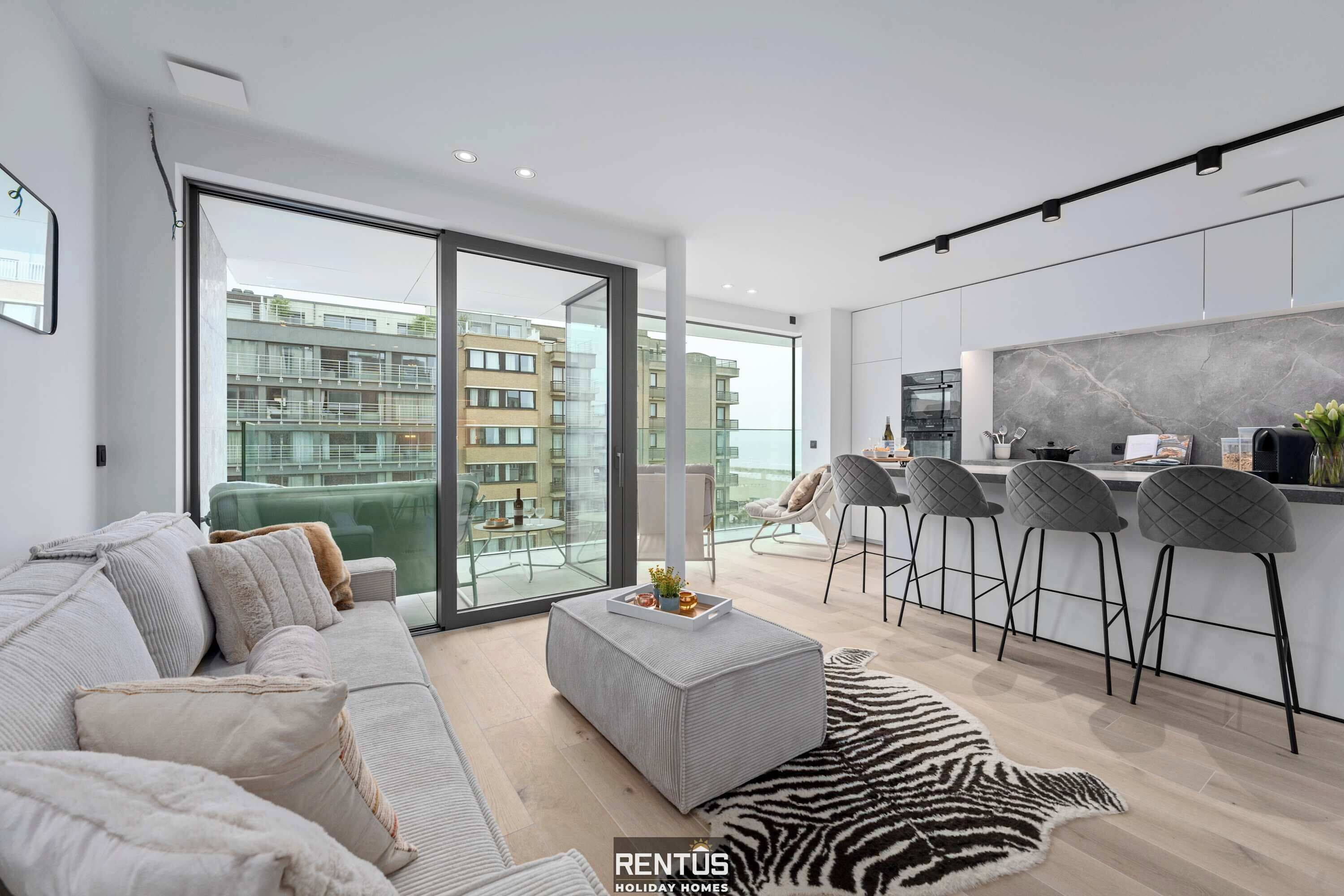 Eine moderne Wohnung mit einer großen Glasschiebetür zu einem Balkon, einer Küche mit Barhocker und einem Sofa mit einer Schale voller Blumen.